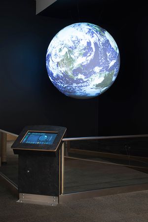 Science On a Sphere hangs in a corner displaying satellite imagery of the Earth. The SOS Public Kiosk is running on a touchscreen in front of the Sphere
