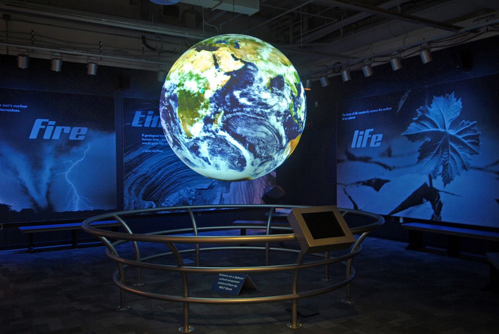 Science On a Sphere hangs at the center of an exhibit room displaying satellite imagery of the Earth. A touchscreen is mounted to the circular guardrail around the Sphere