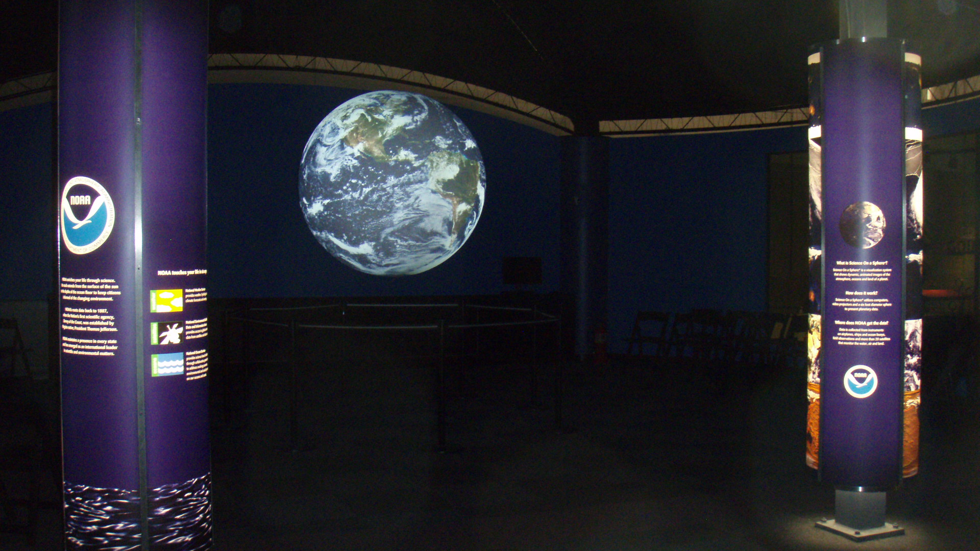Science On a Sphere displays satellite imagery of Earth in an empty room. In the foreground are two pillars showing the NOAA logo and some explanatory text