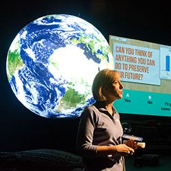 A presenter stands in front of Science On a Sphere. The dark area of the Sphere is not visible because it is on the side opposite the audience