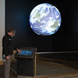 A person stands that SOS Public Kiosk touchscreen in front of a Science On a Sphere that hangs in a corner