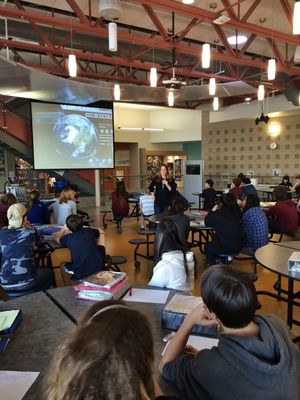 Students sit around tables in a cafeteria looking at an image of the earth projected on a screen while Hilary stands in front of the screen holding a microphone