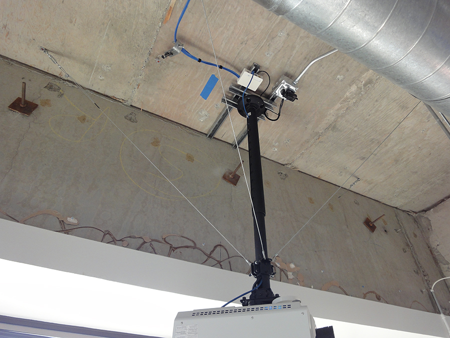 A projector is mounted to a long pole attached to the ceiling by two pairs of metal tracks placed at 90 degrees to one another. Three stabilizing wires extend from the pole where it attaches to the projector up to the ceiling