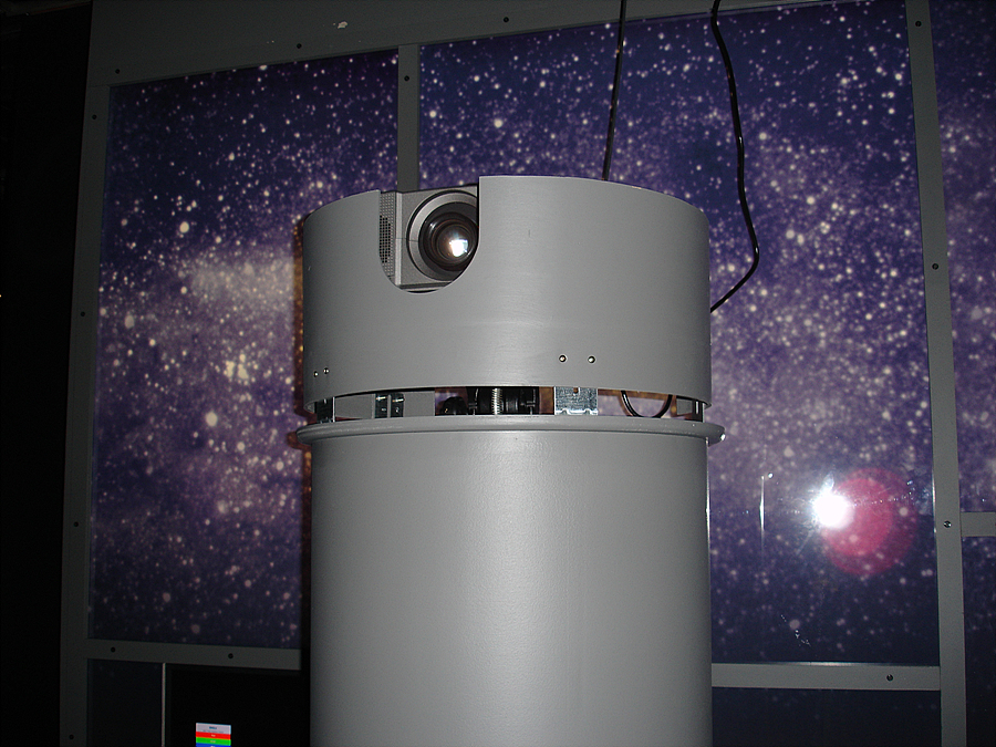 A projector lens is just visible inside a u-shaped cut out at the top of a gray cylinder that surrounds the stand on which the projector is mounted