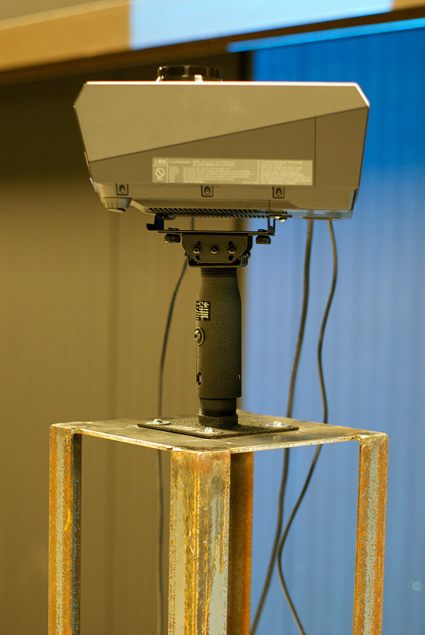 A projector is mounted atop a square stand made of solid, flat metal