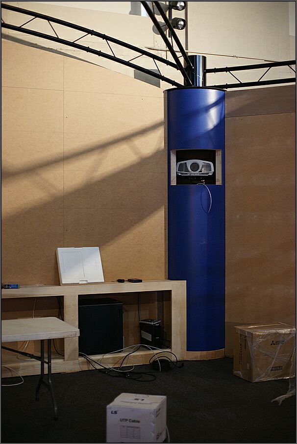 A projector is visible inside a window cut into a blue cylinder that hides the floor stand on which it is mounted