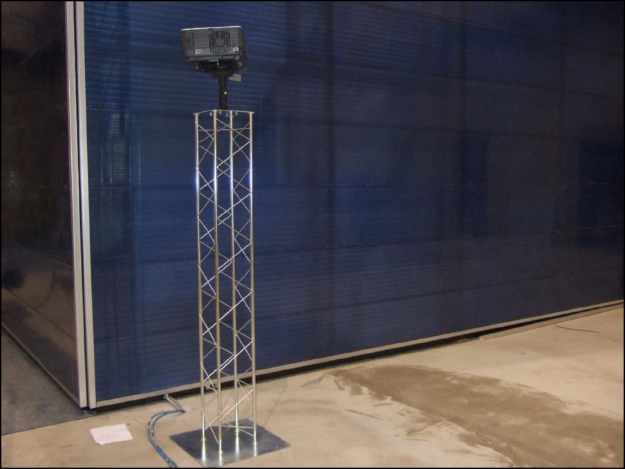 A projector is mounted atop a metal stand made of four poles with criss-crossing metal rods to stabilize it