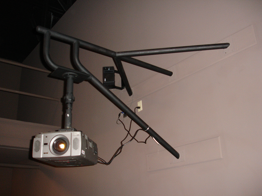 A projector is mounted to a vertical metal rod attached to a complex frame created of welded and bent black metal rods. The frame attaches to the wall at four different points