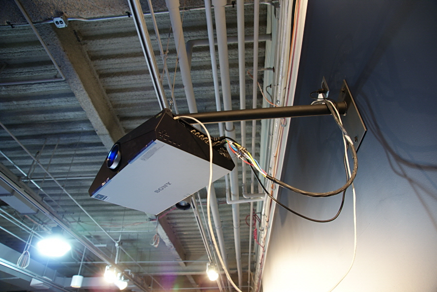 A projector is mounted to a short pole that extends down from a longer black pole attached to the wall