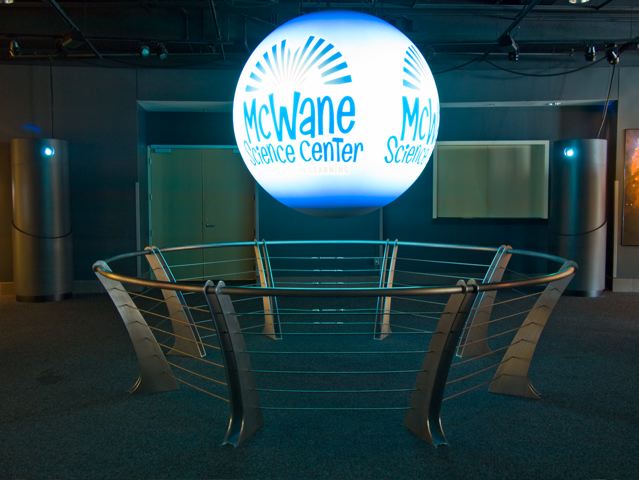 A circular metal rail extends up and out from the floor surrounding Science On a Sphere as it displays the McWane Science Center logo