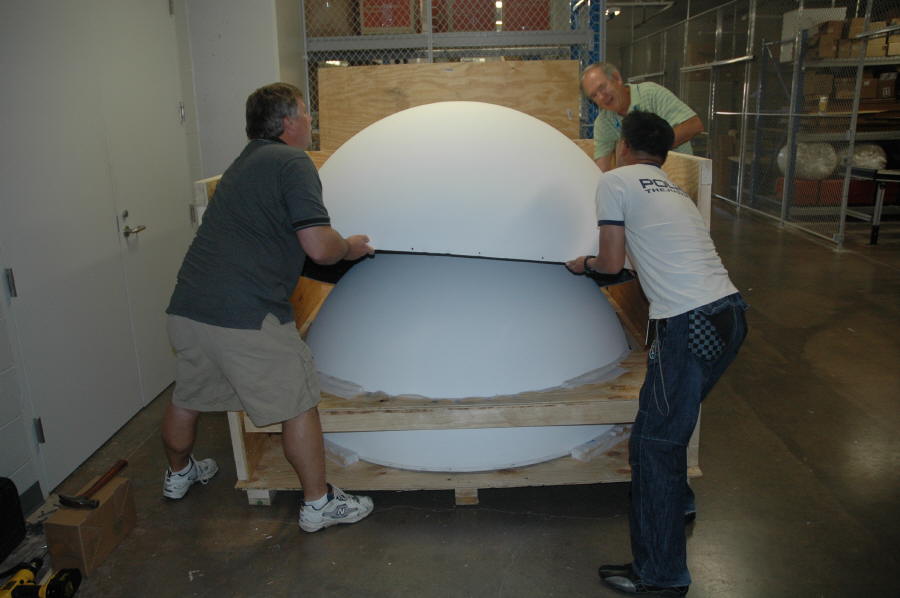 Three people lift the upper half of a Science On a Sphere out of its crate
