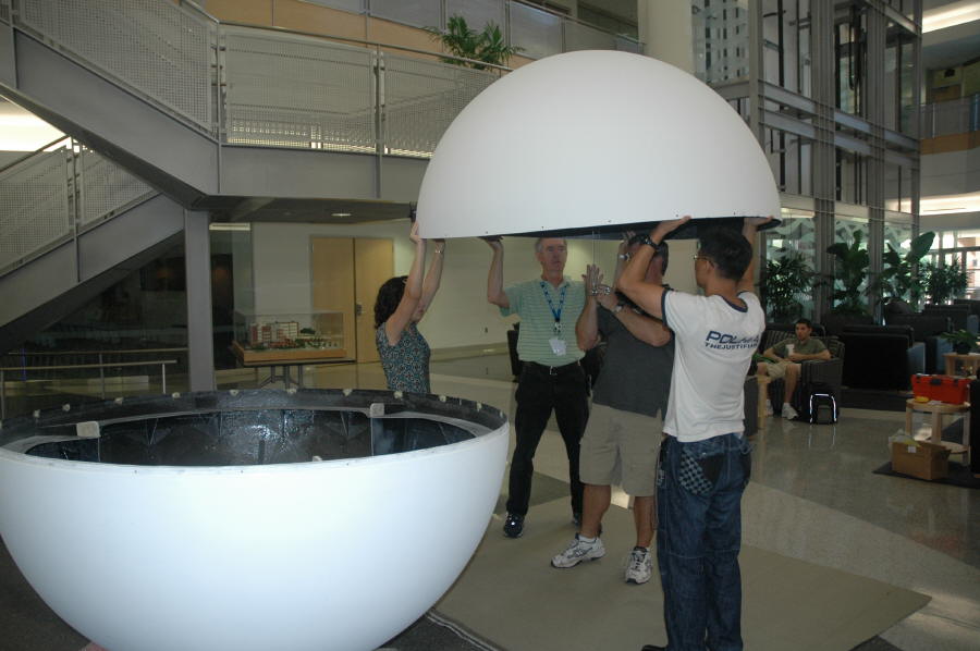 The bottom half of a Science On a Sphere rests on the floor as four people lift the top half and prepare to set it on top