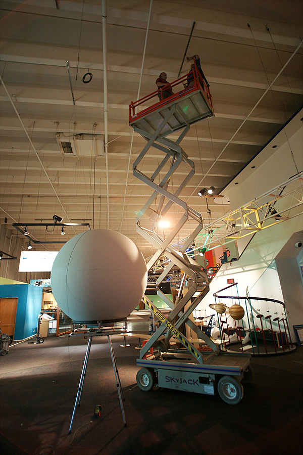 An assembled Science On a Sphere sits on a stand as two people in a cherry picker examine the mounts in the ceiling from which it will be hung