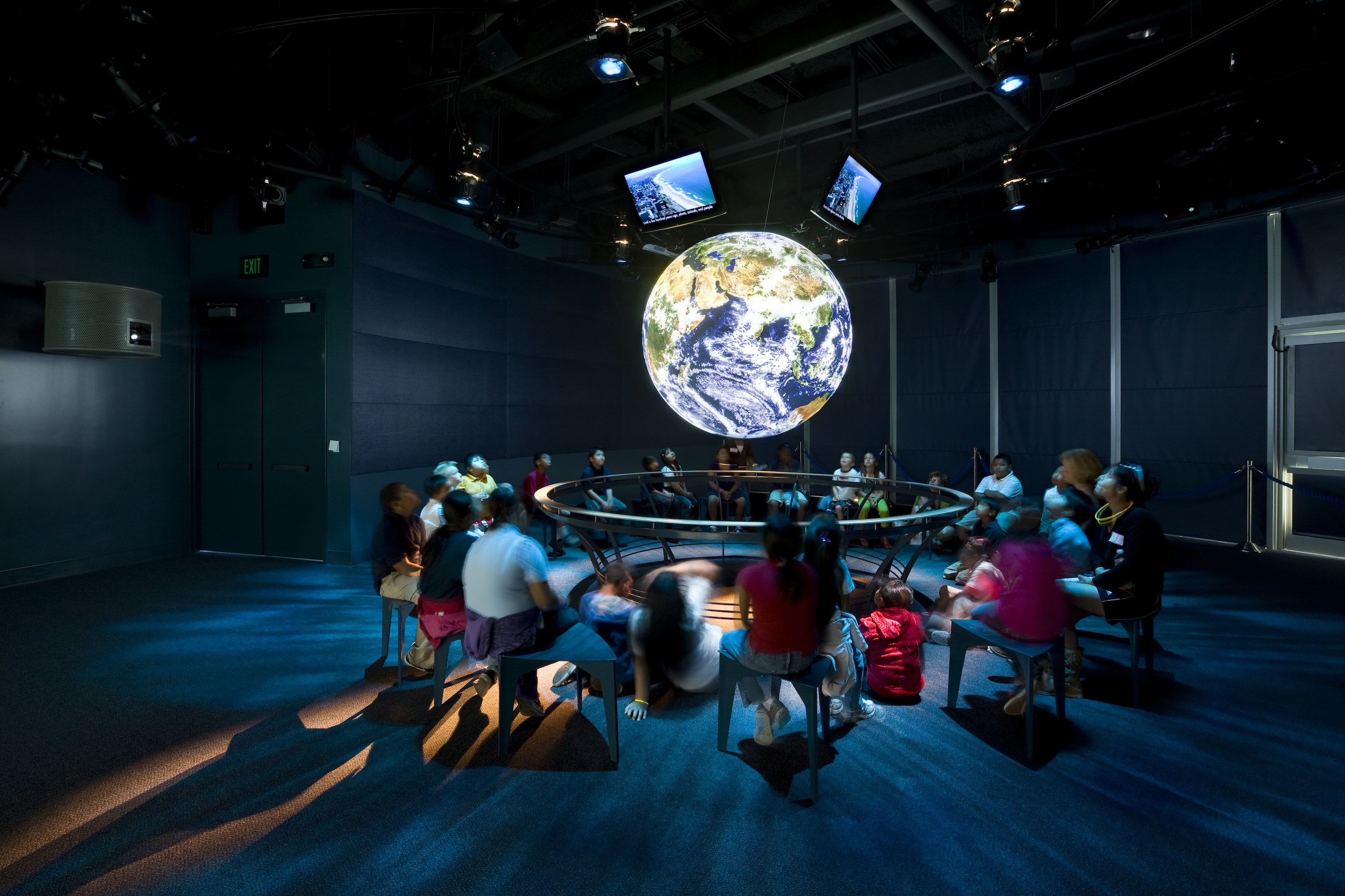 A group of people sit on stools or on the floor surrounding Science On a Sphere in a darkened theater. Four monitors are mounted above the Sphere facing outward to display additional information for the audience