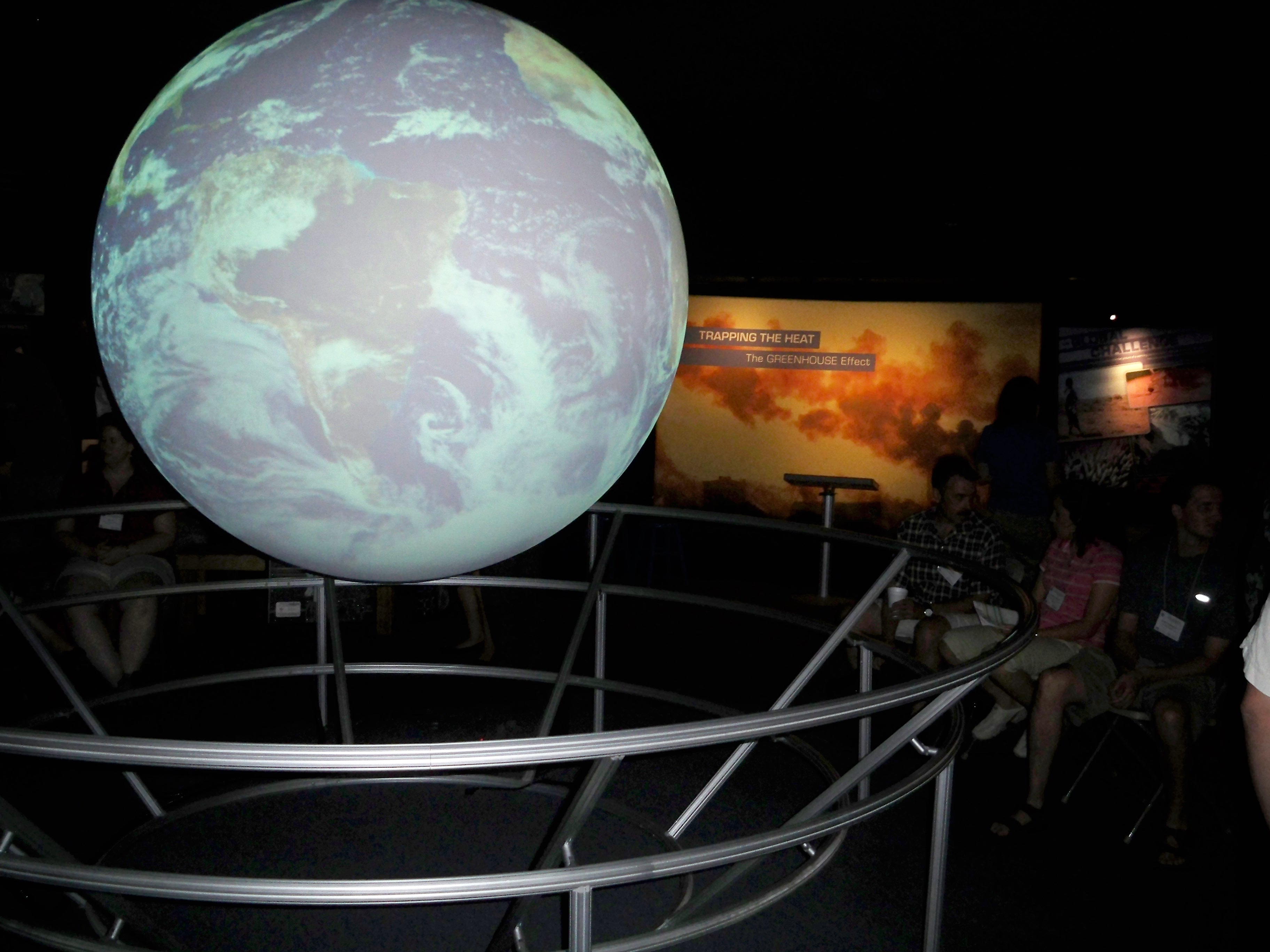 A group of people sit in folding chairs watching Science On a Sphere in a darkened room. Behind the Sphere another exhibit titled 'Trapping the Heat: The Greenhouse Effect' is visible