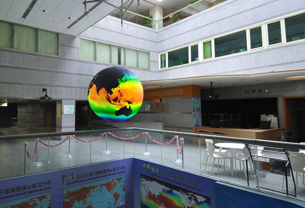 Science On a Sphere hangs in an atrium, surrounded by a balcony. It is displaying ocean data in hues of red, yellow, green, and blue; the continents are black