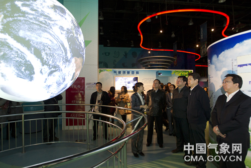 A group of people in a well-lit exhibit hall stand and watch Science On a Sphere displaying satellite imagery of earth