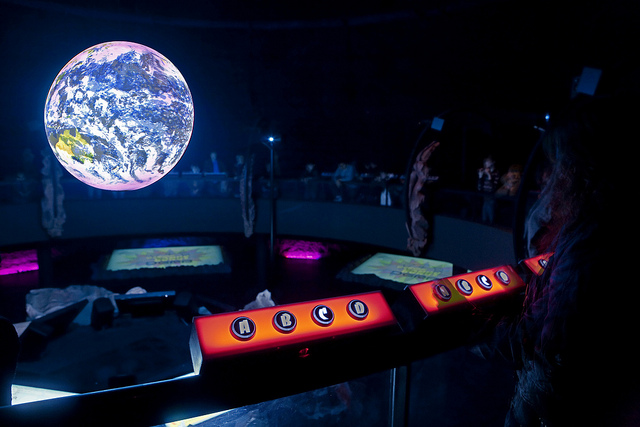 Science On a Sphere glows in the background of a dark room. In the foreground, several groups of four buttons labelled A, B, C, and D are mounted to the railing in rectangles that glows orange