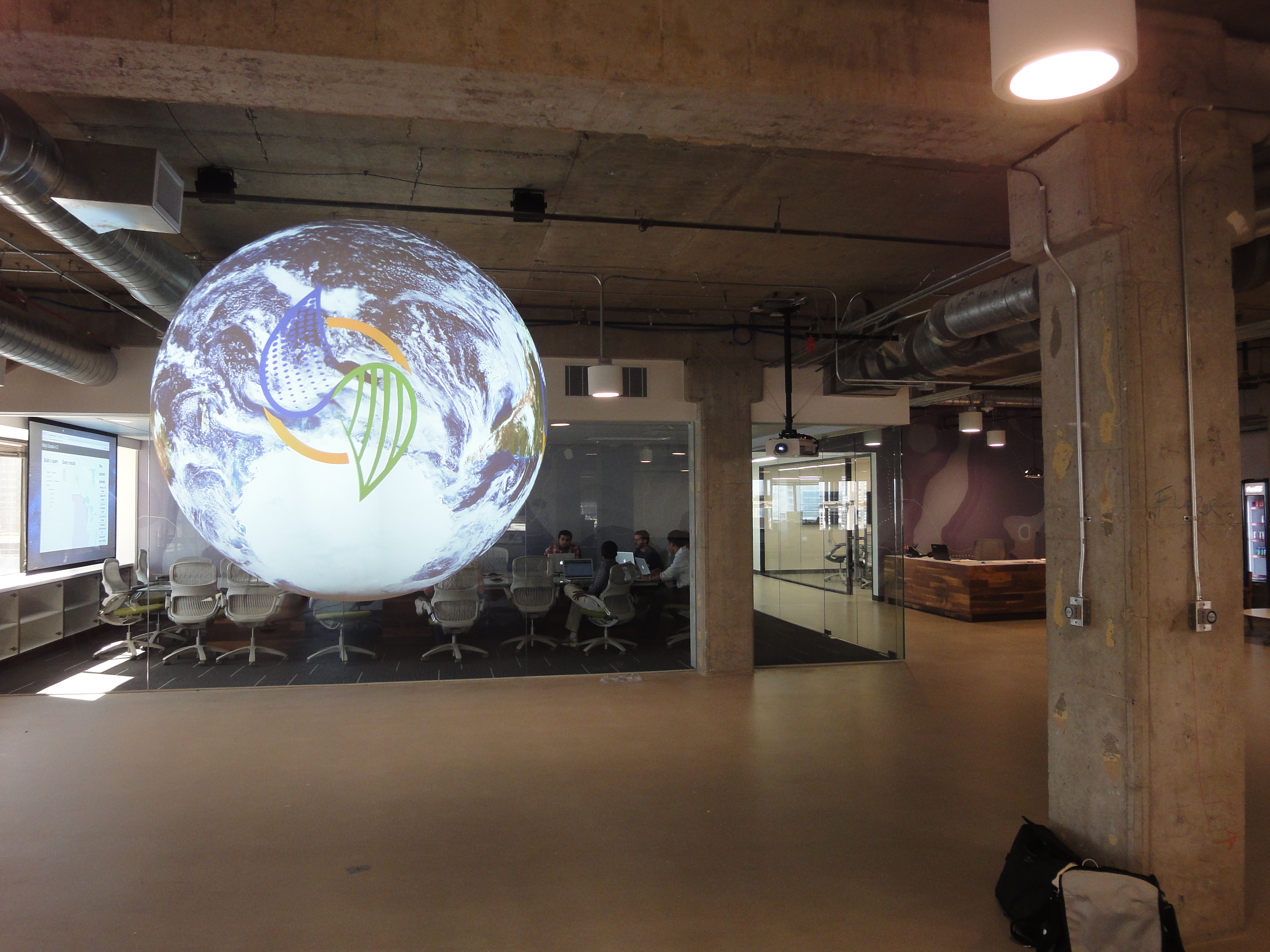 Science On a Sphere displays the Climate Corp logo over a satellite image of Earth in concrete lobby. In the background some people sit around a large conference table in a room with glass walls