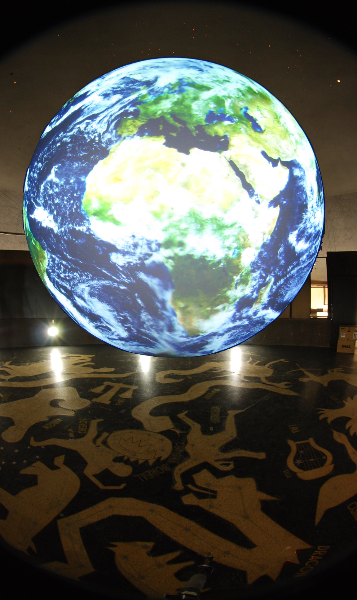 Science On a Sphere displays satellite imagery of Earth in an empty room. On the floor of the room are images of constellations.