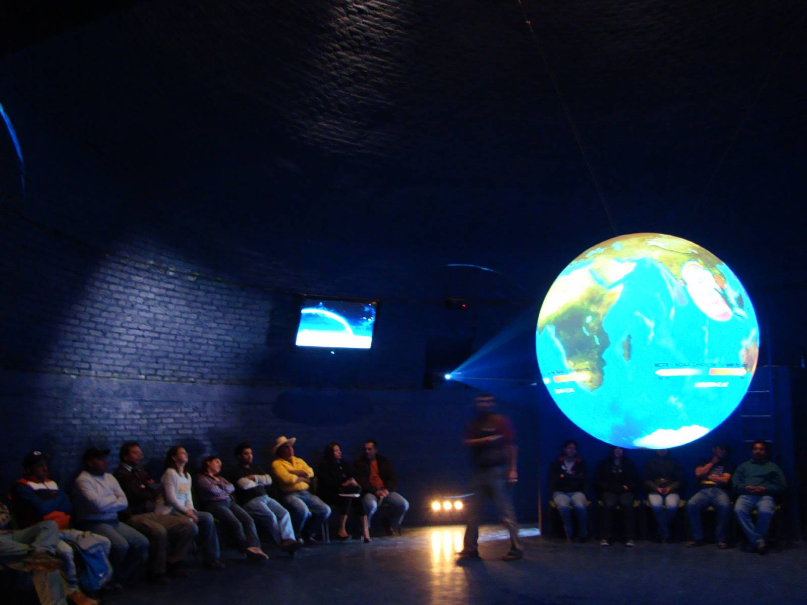 An audience sits against a brick wall in a circular room watching a presentation on Science On a Sphere