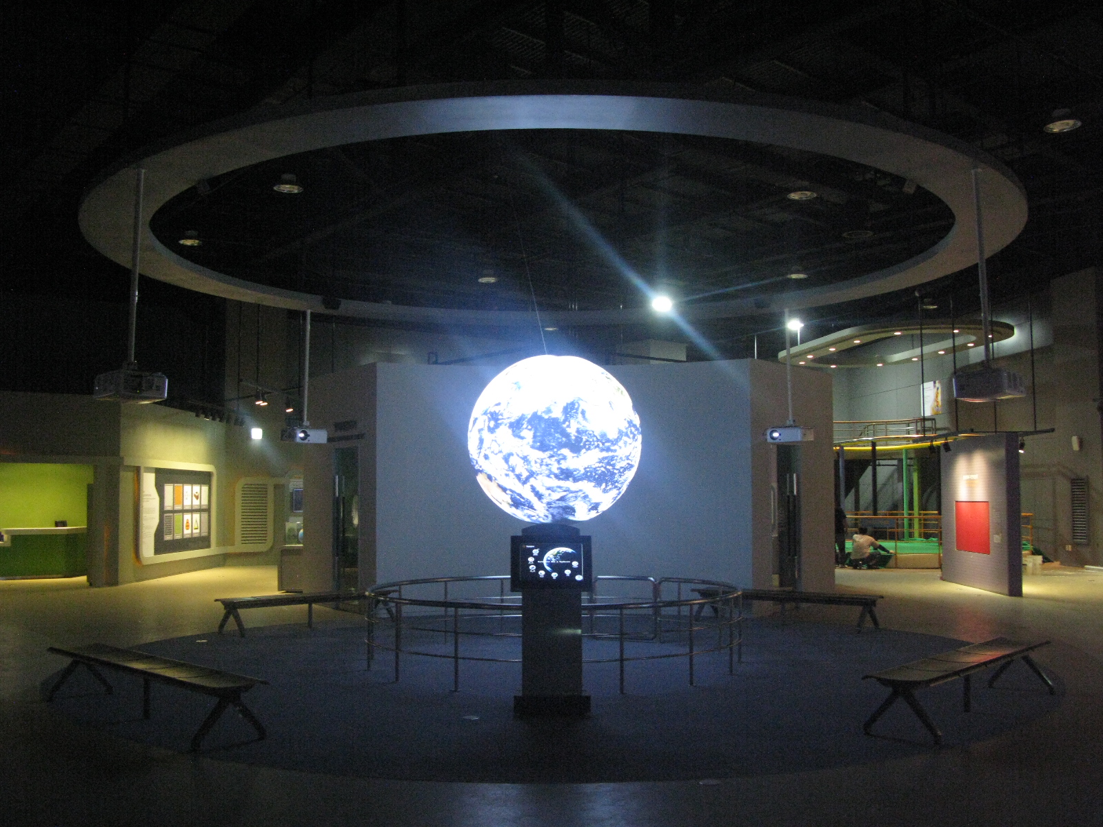 Science On a Sphere hangs in a large exhibit space. In the foreground is a touchscreen, and the Sphere is surrounded by four benches. Other exhibits are visible in the background