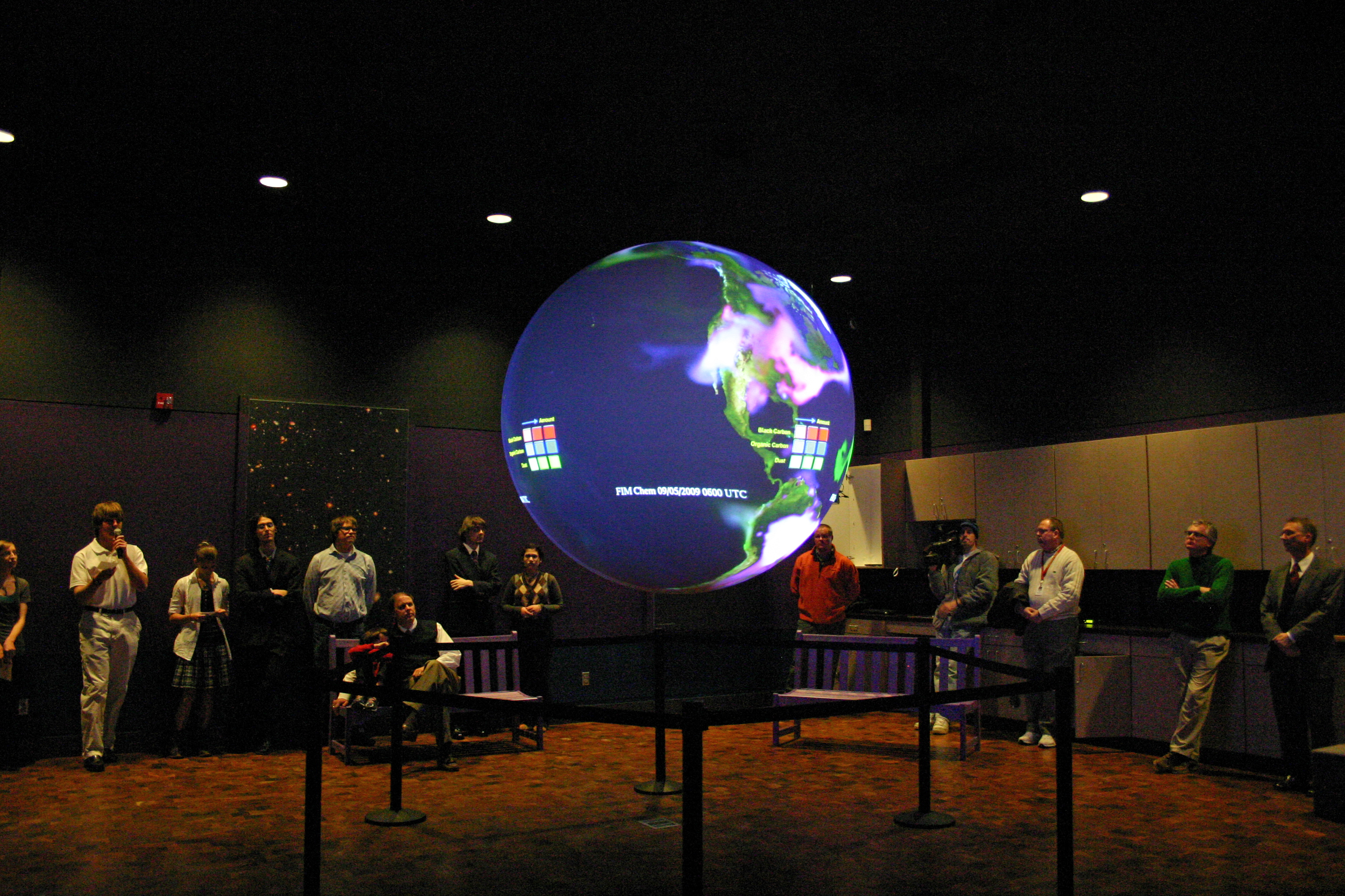 A group of adults stand at the edges of a room watching Science On a Sphere