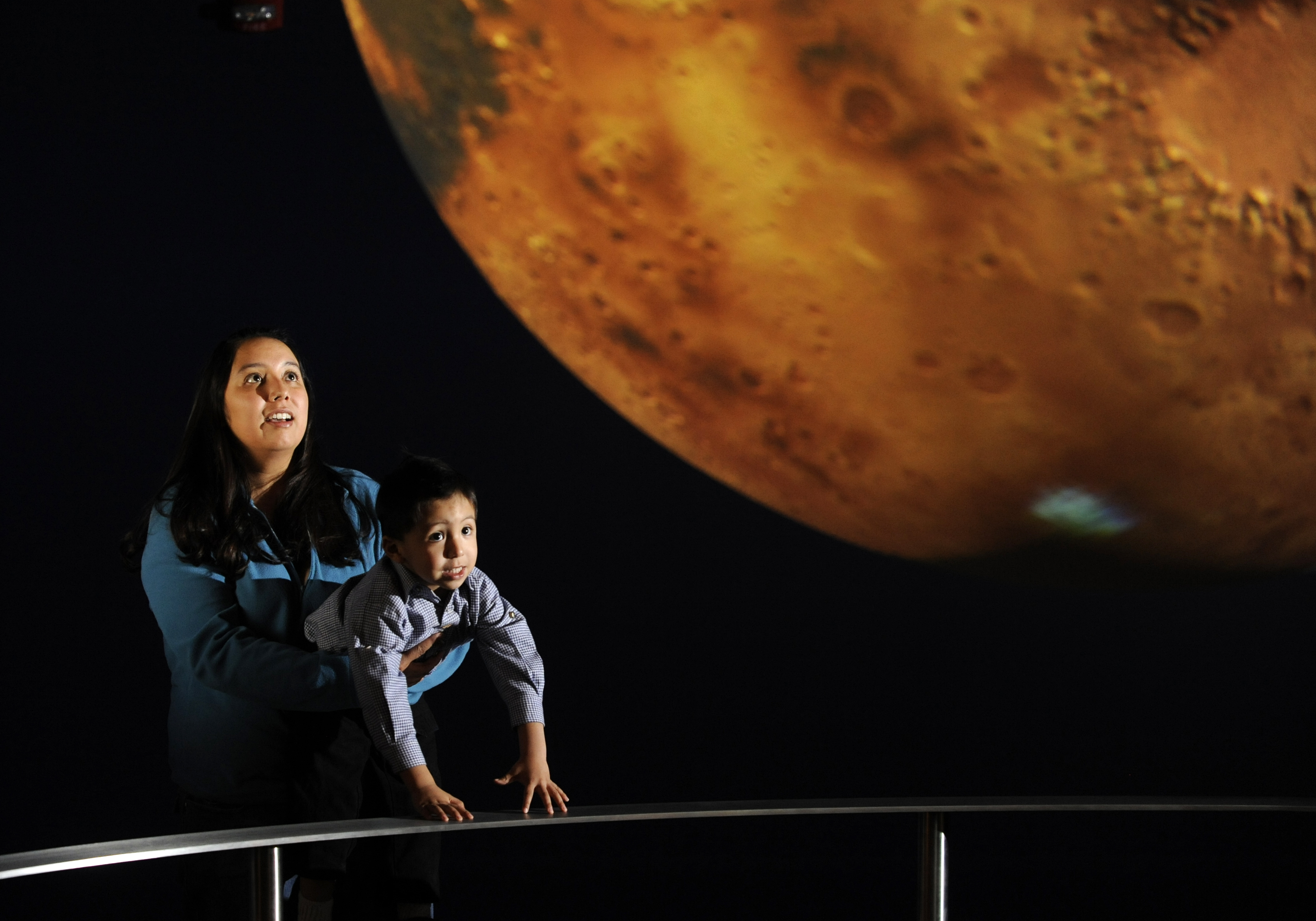 A woman holds a child up over the railing as they look at Science On a Sphere displaying imagery of Mars