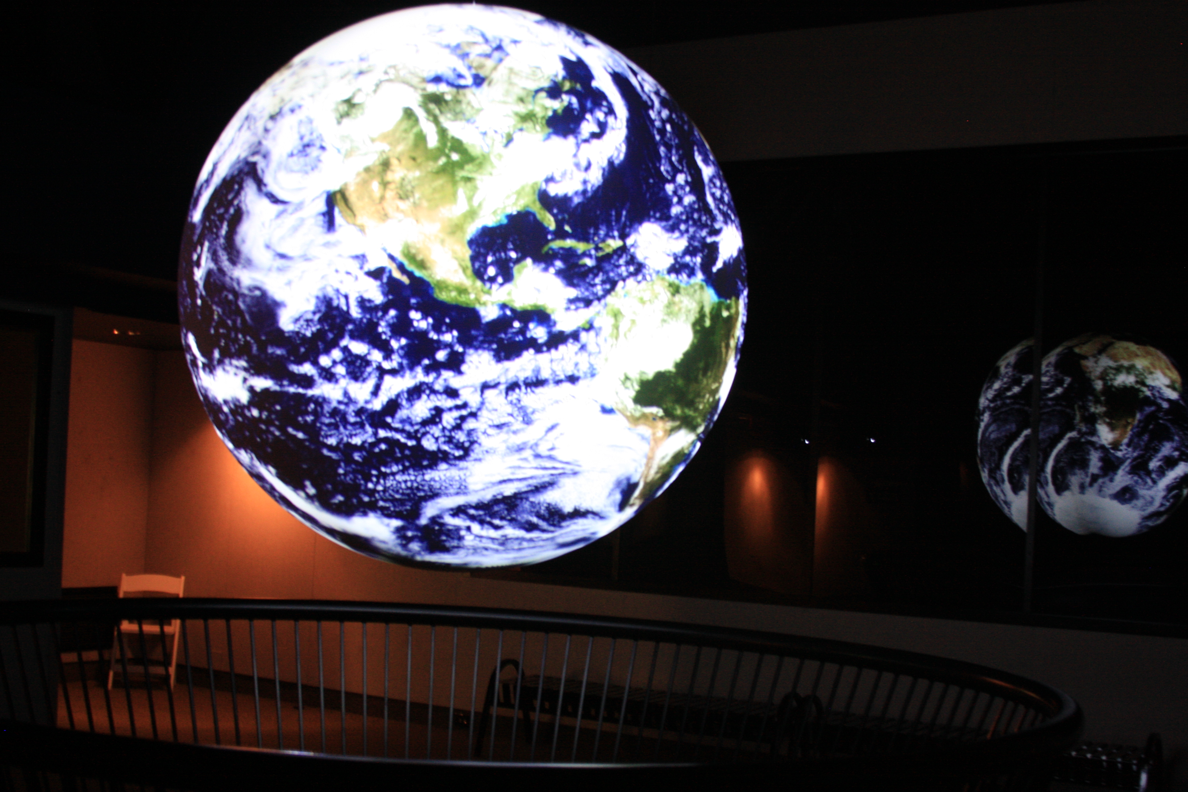 Science On a Sphere displays satellite imagery of the Earth in an empty theater. A reflection of the Sphere is visible in some glass in the background