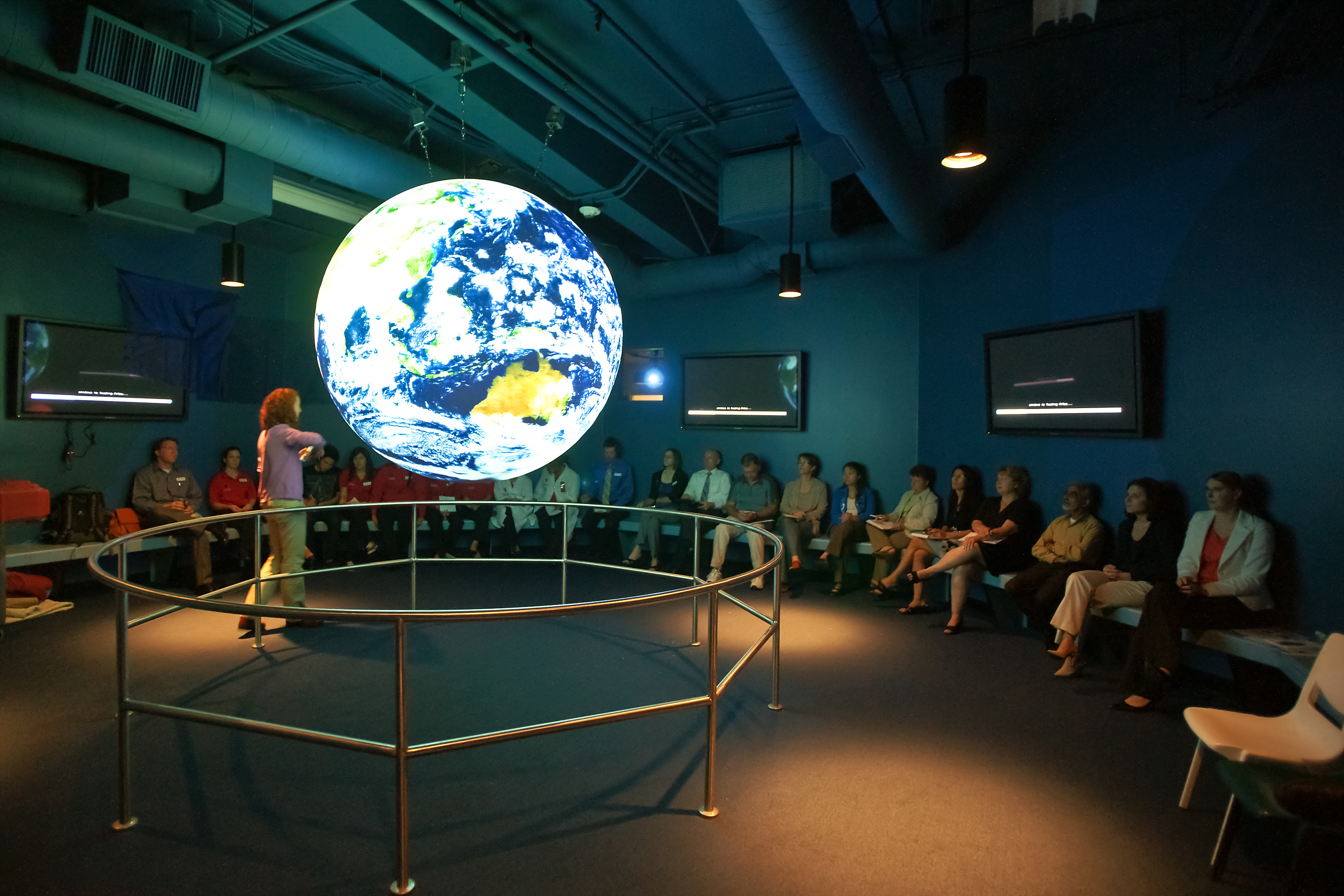 A group of adults sit on benches lining the walls surrounding Science On a Sphere watching a presentation