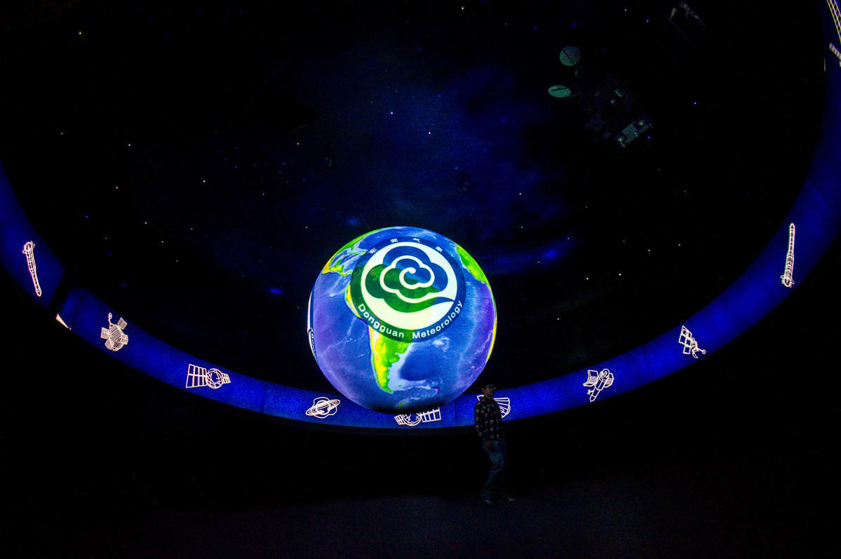 The camera looks up at an angle from the floor at Science On a Sphere displaying the logo for the Dongguan Meteorology and Astronomy Museum overlaid atop an image of Earth. In the background white line drawings of satellites against blue backgrounds adorn the walls surrounding the Sphere