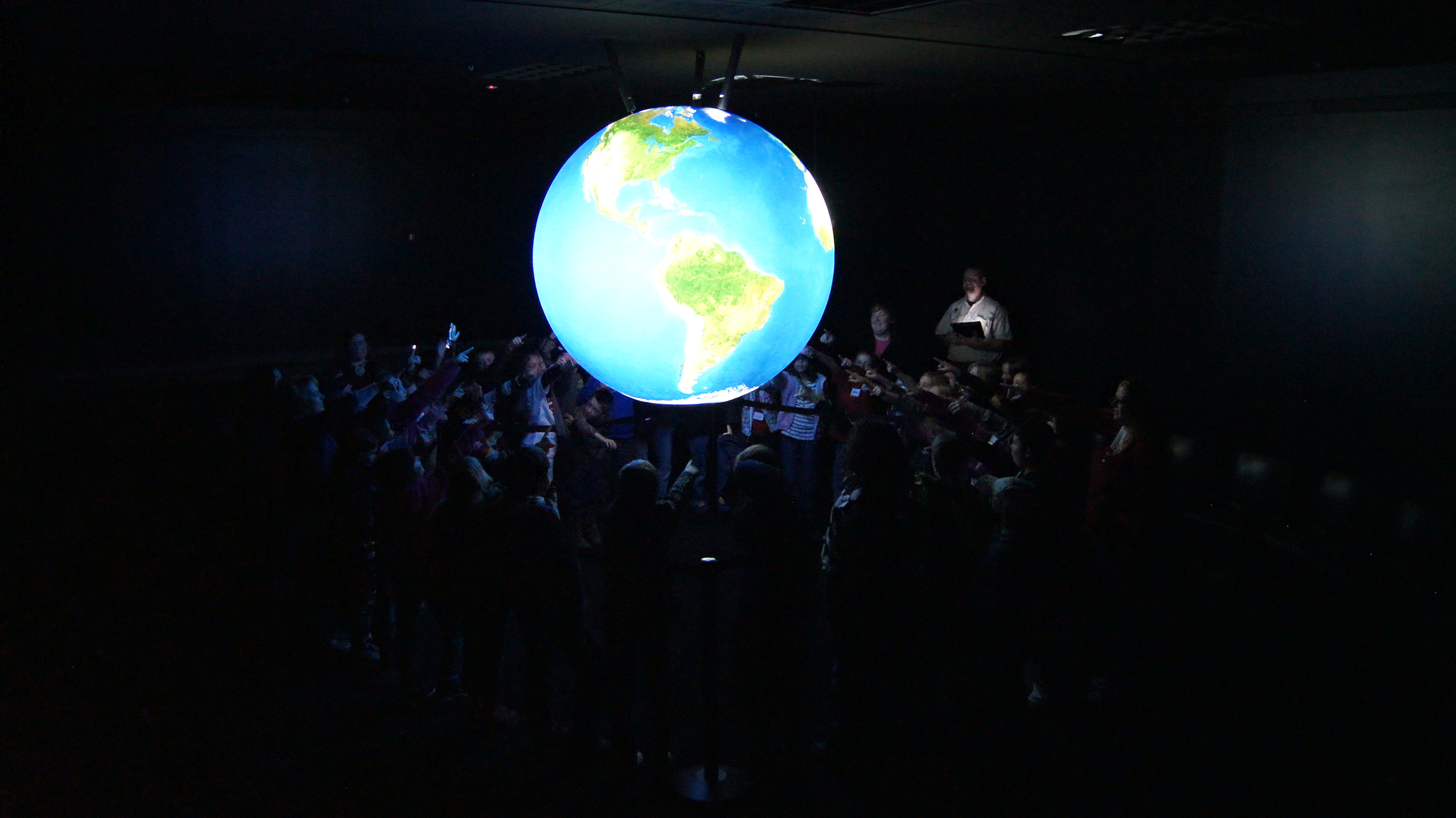 A group of school children stand around Science On a Sphere, pointing at it. They are illuminated only by the light reflected from the Sphere