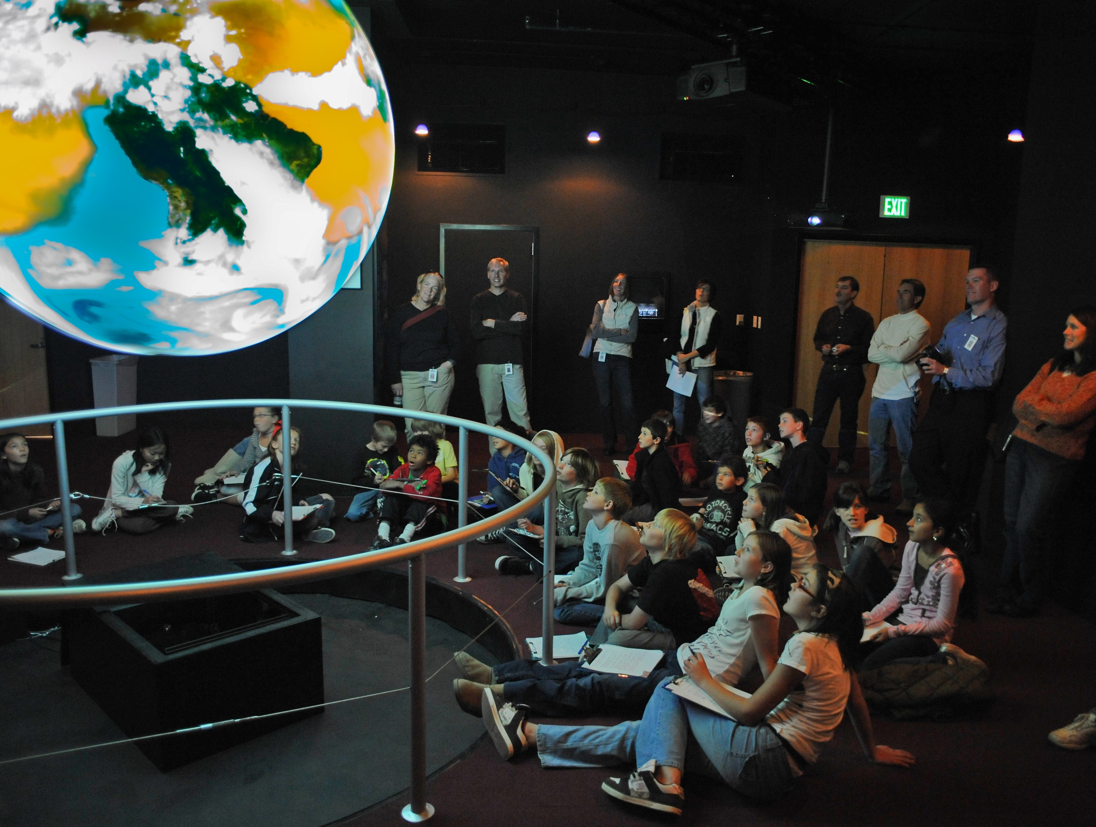 A group of children sit on the floor around Science On a Sphere as some adults stand behind them watching a presentation on Science On a Sphere