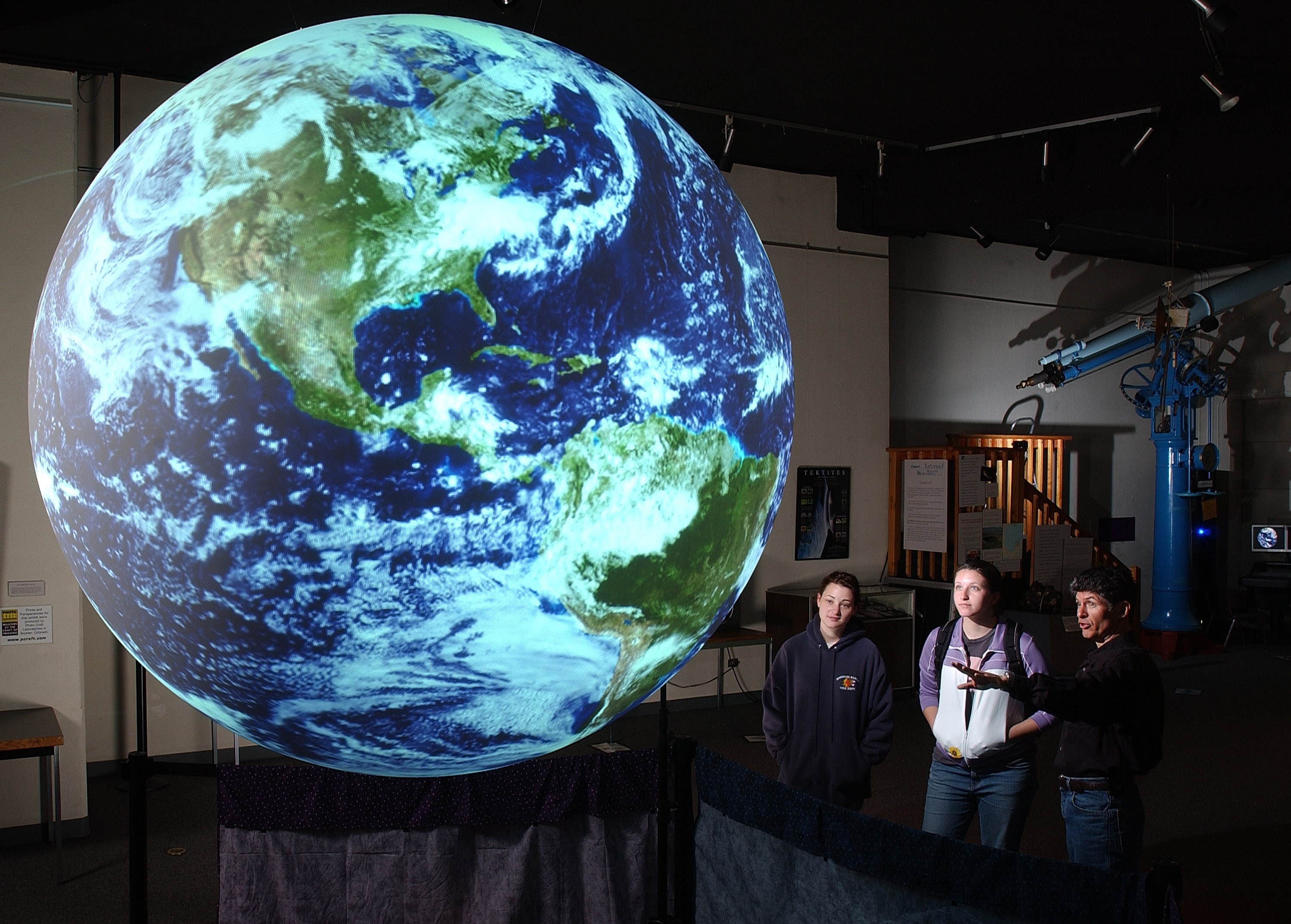 Three people stand in front of Science On a Sphere, one of them is gesturing towards the Sphere and speaking. In the background, a telescope is visible