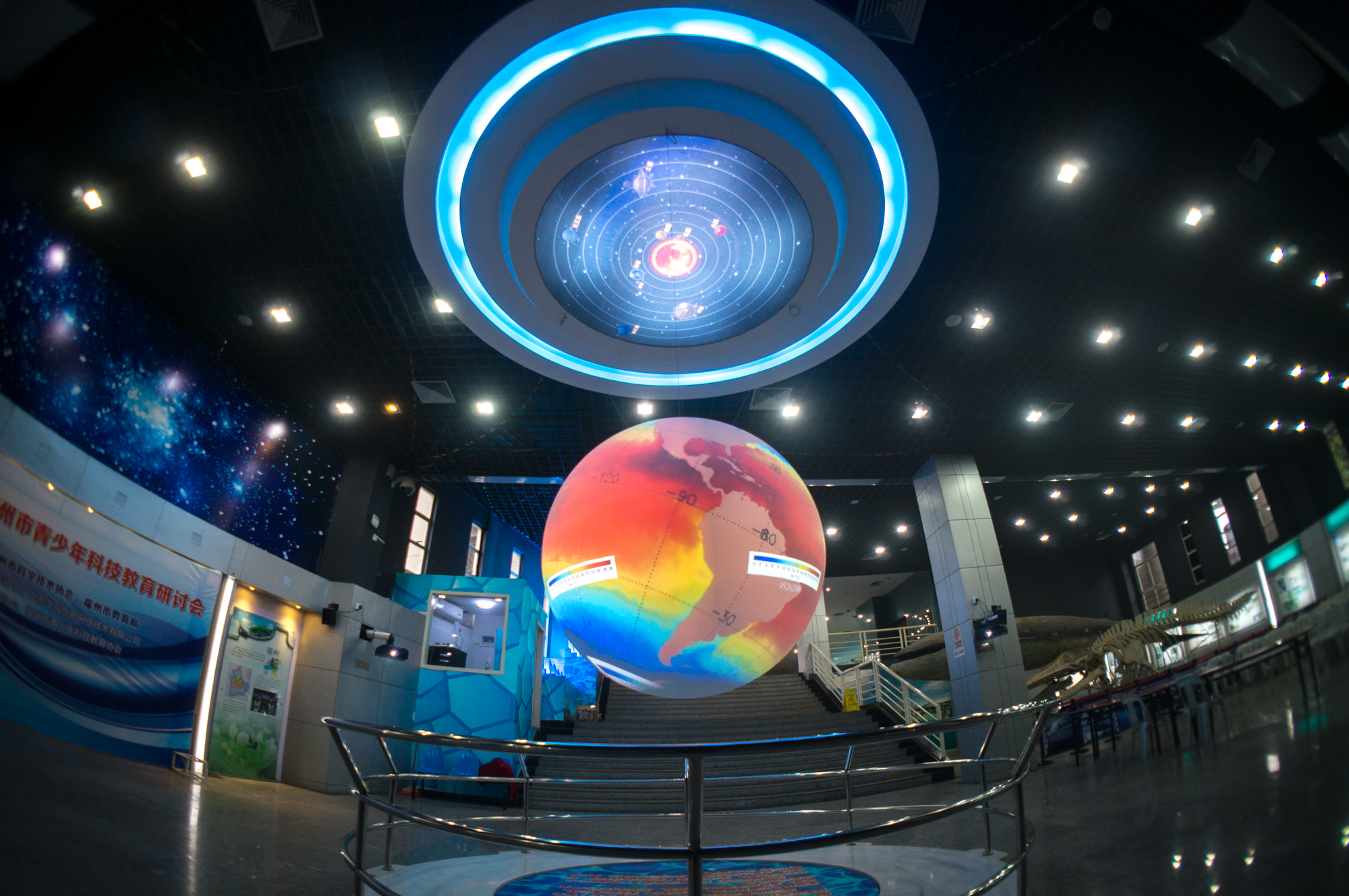 Science On a Sphere hangs in a large, well-lit exhibit space. The circular fixture from which it hangs glows blue and displays a diagram of the solar system. In the background, a model and a skeleton of a whale are both visible