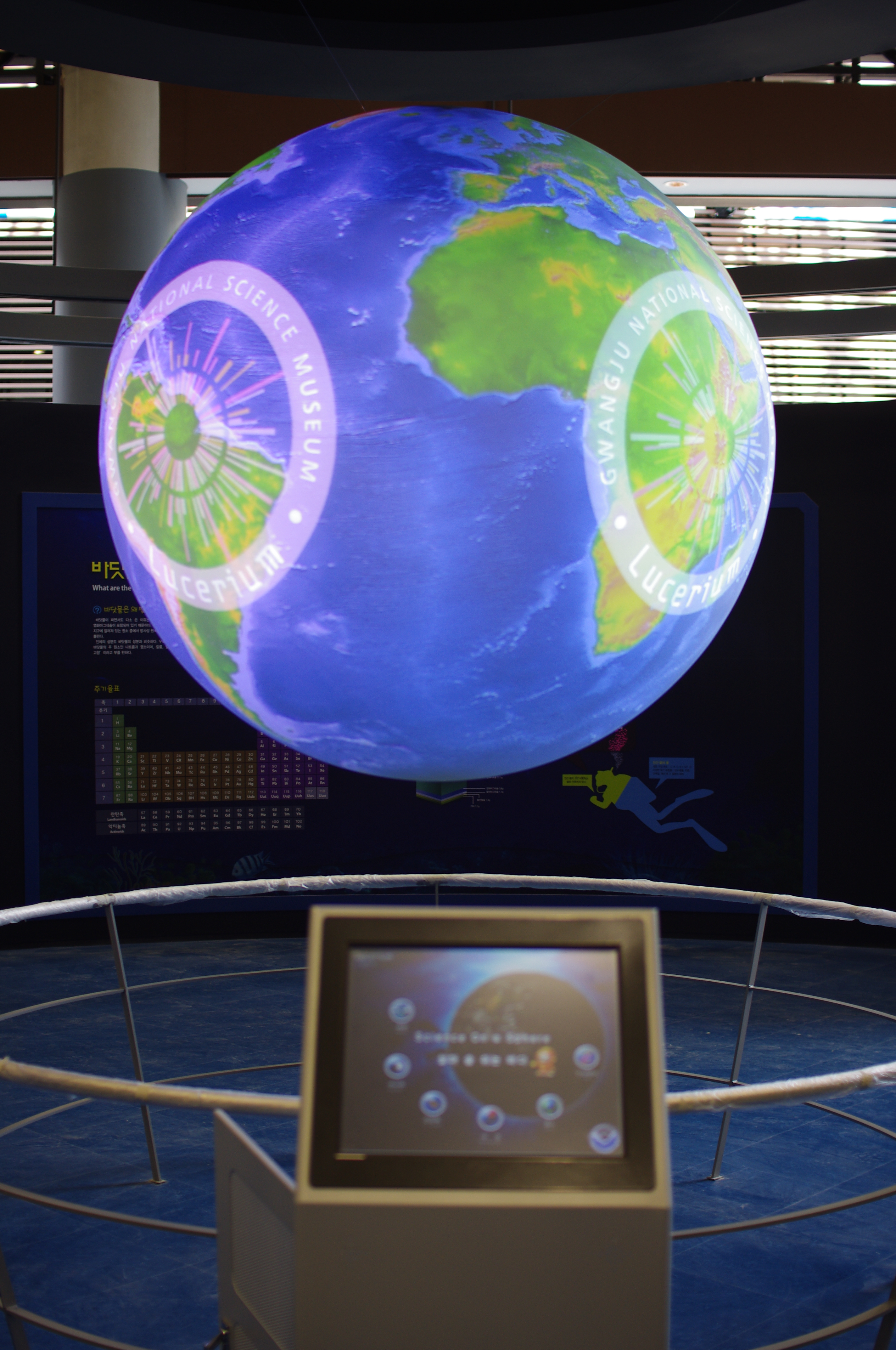 Science On a Sphere displays the Gwangju National Science Museum logo atop an image of Earth. A touchscreen for controlling the Sphere is visible in the foregorund