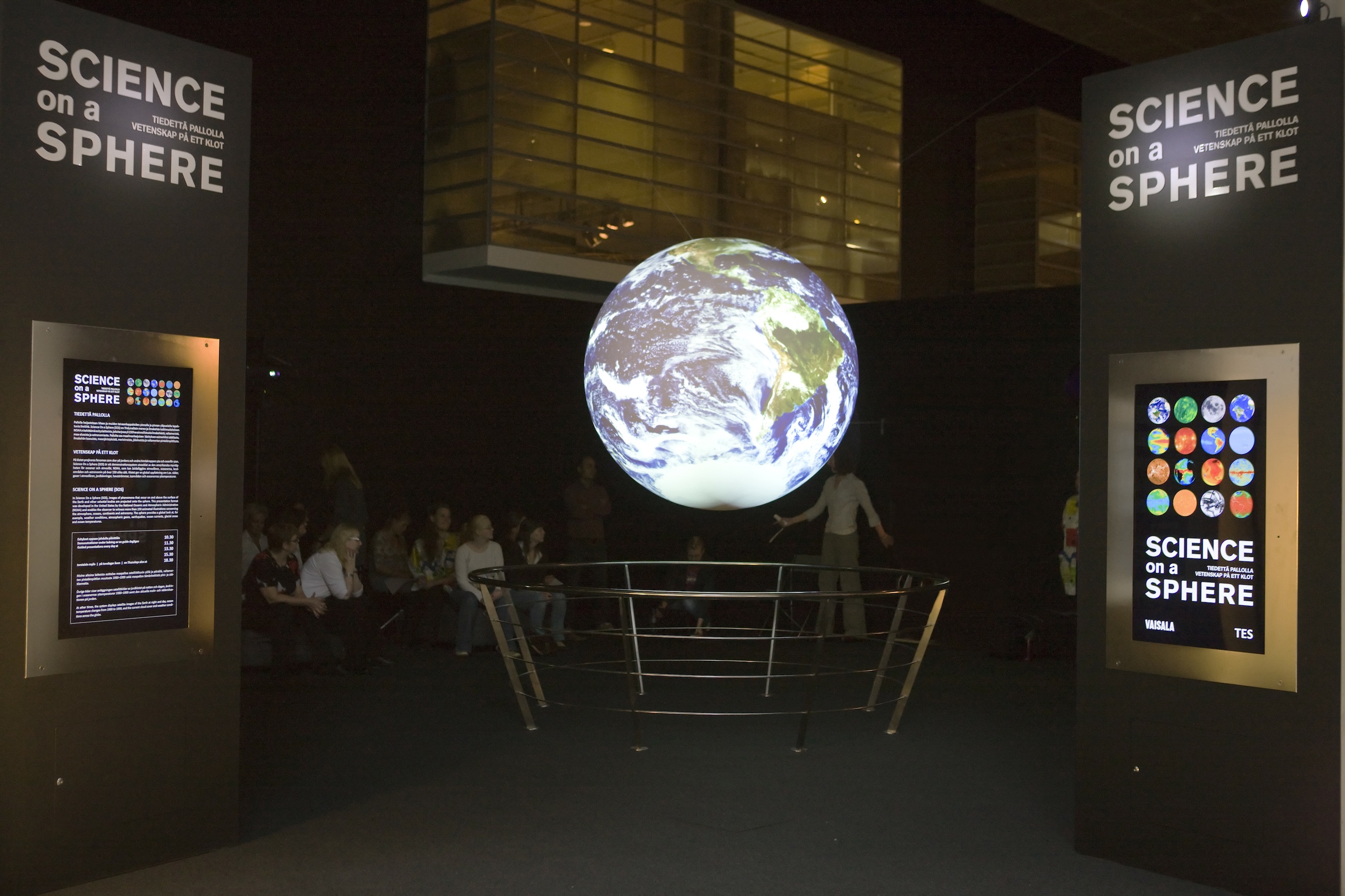 A group of people sit, watching a presentation on Science On a Sphere. Two signs with vertical monitors embedded in them frame the Sphere in the foreground