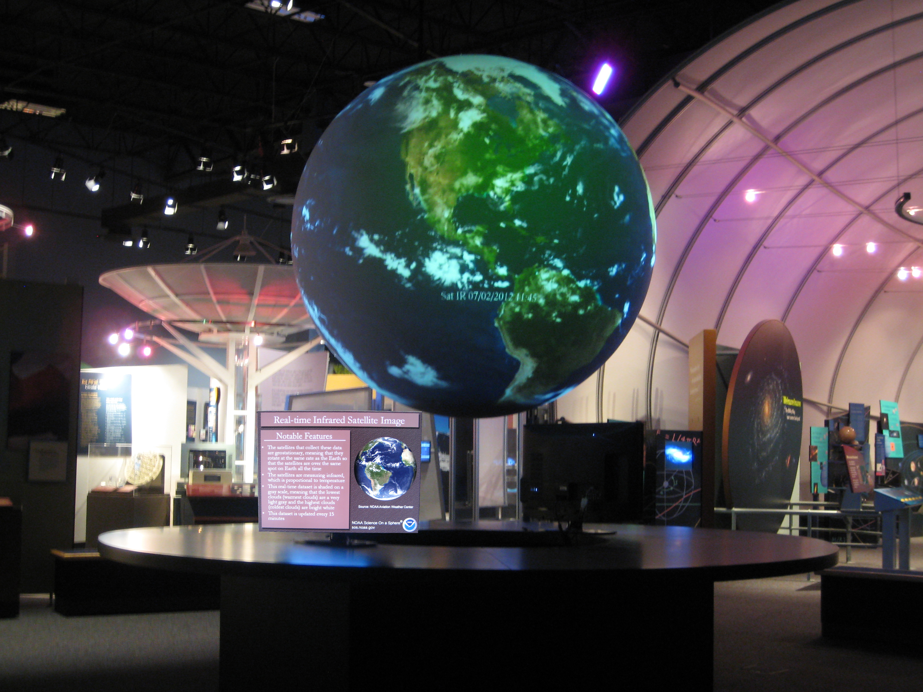 Science On a Sphere hangs in an empty exhibit space over a round table. Two monitors are attached to the table displaying information about the dataset (Real-time Infrared Satellite Image) currently displayed on the Sphere. More exhibits are visible in the background