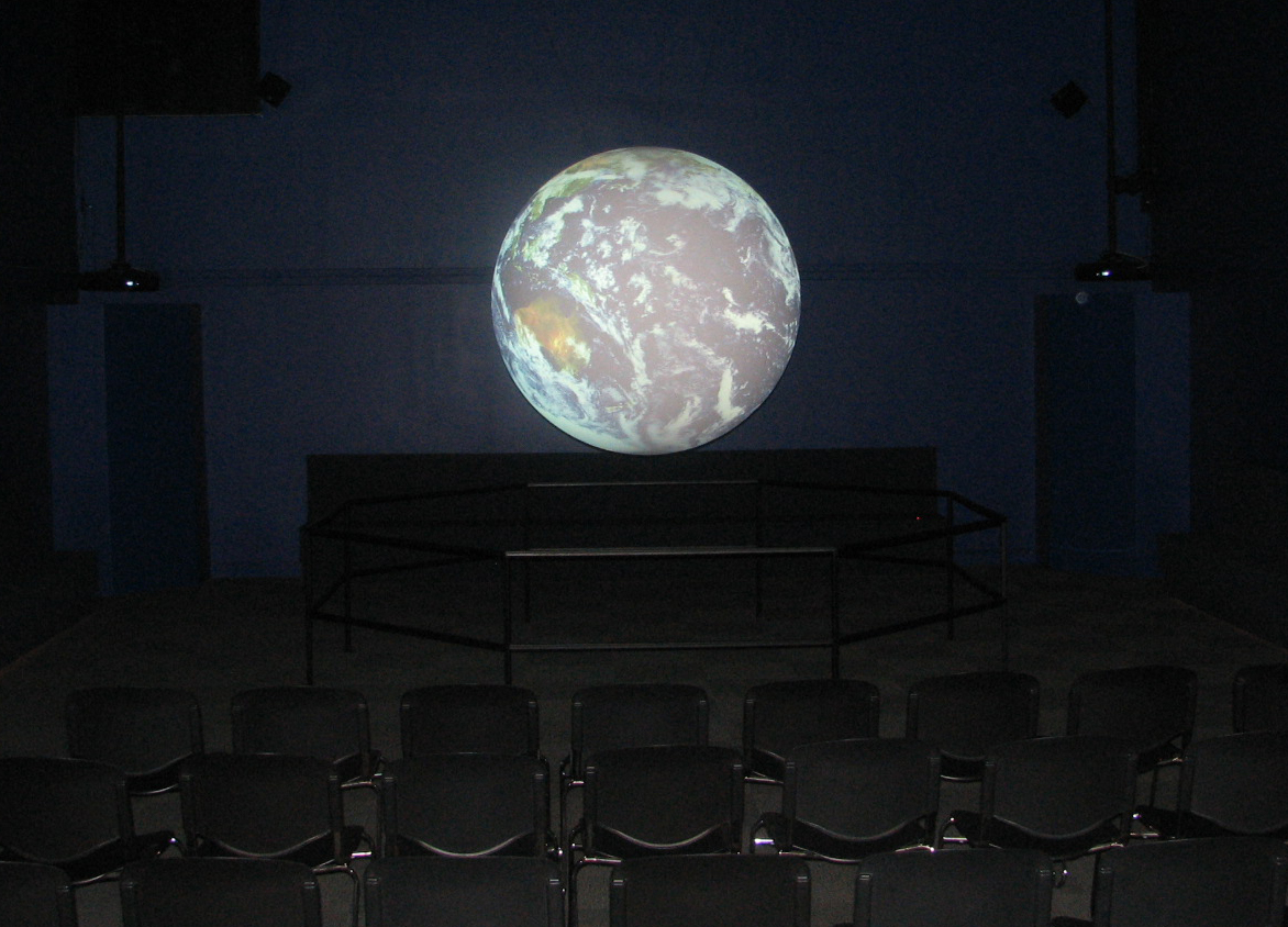 Science On a Sphere displays satellite imagery of Earth in an empty theater. Rows of black chairs are visible in the foreground