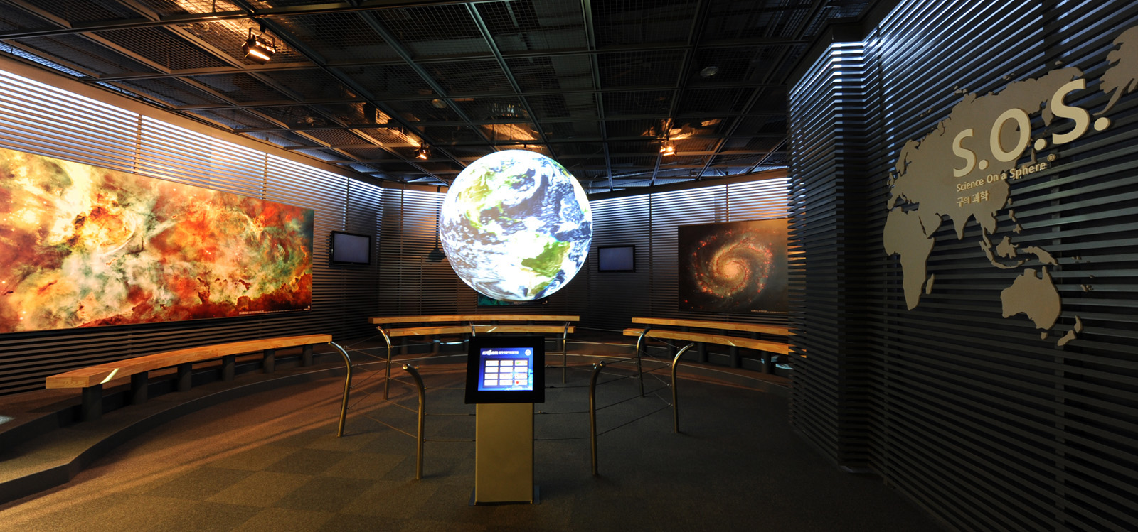 Science On a Sphere displays satellite imagery of Earth in an empty room. A touchscreen sits in front of the Sphere, and behind it are several benches. Photos of a galaxy and a nebula adorn the walls in the background
