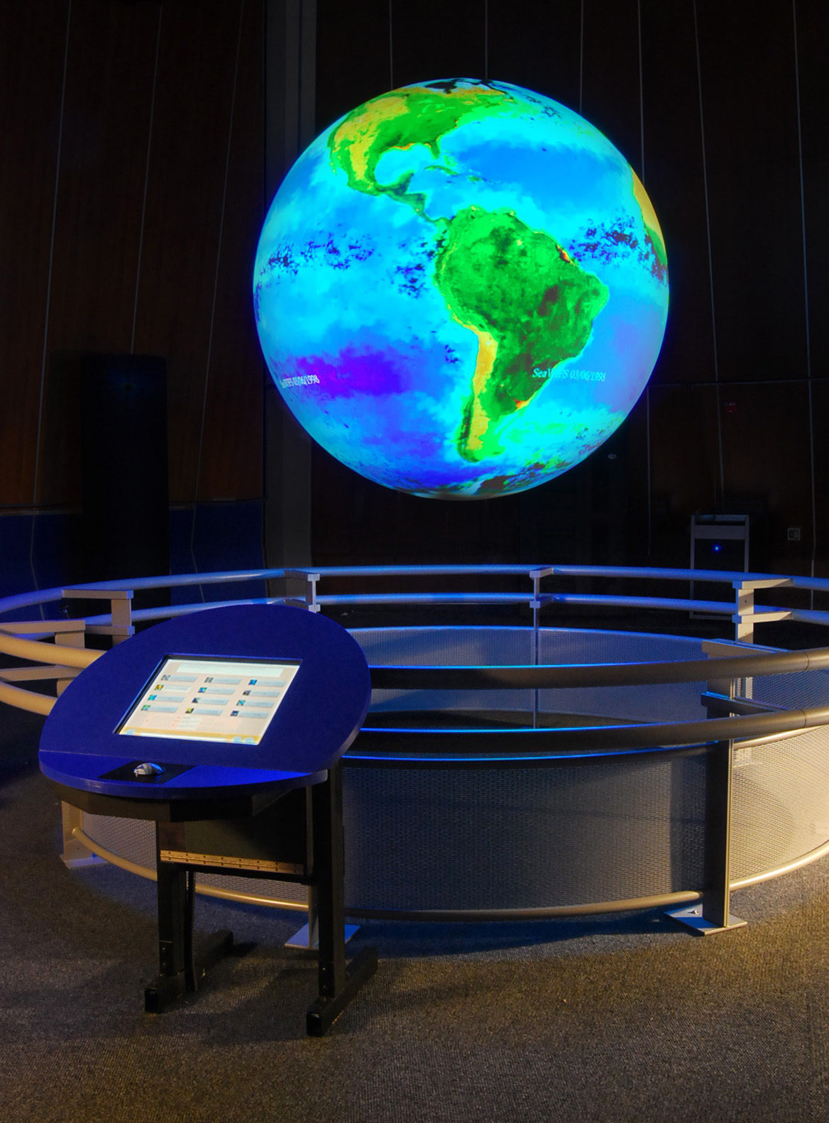 Science On a Sphere hangs in an empty room displaying an image of Earth that depicts North and South America in hues of green, yellow, and red, and the oceans in hues of blue and purple. A touchscreen is visible in the foreground