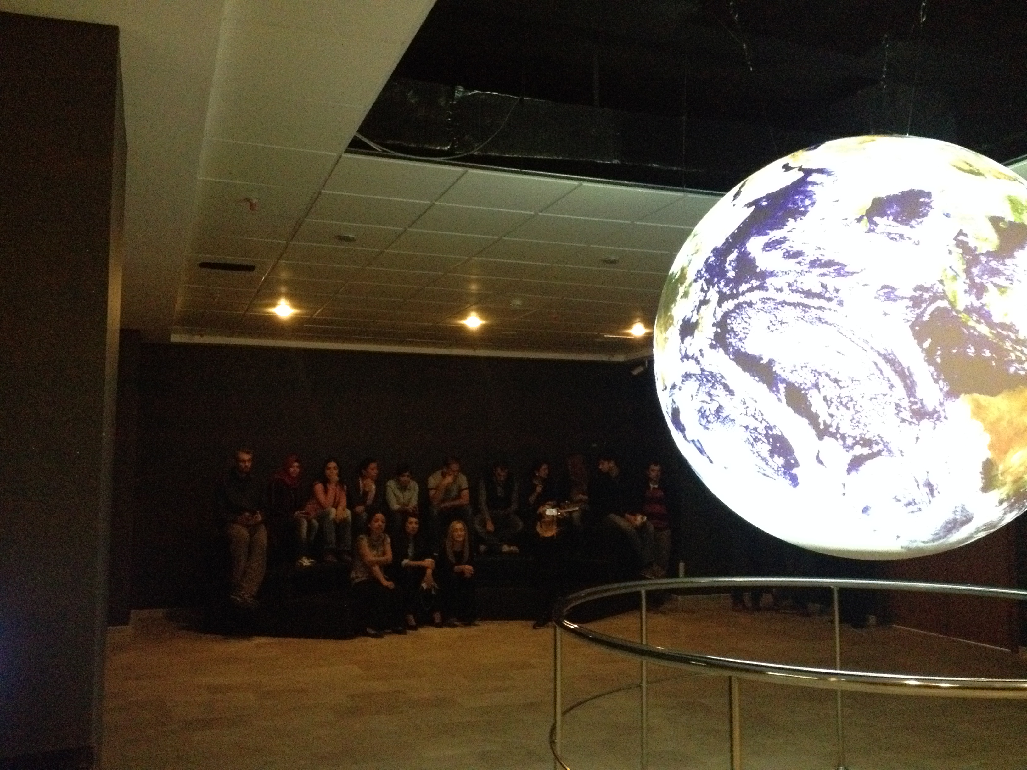 A group of people sit on terraced benches in a theater watching Science On a Sphere