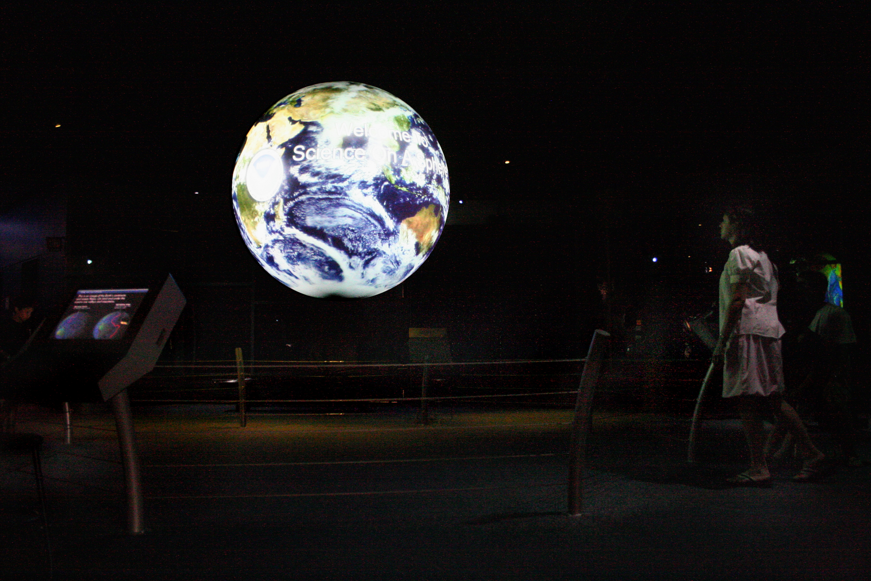 Science On a Sphere displays satellite imagery of Earth in a darkened room. In the foreground, a small screen is attached to the rail displaying additional information. Two museum patrons are standing at the right of the image watching the Sphere