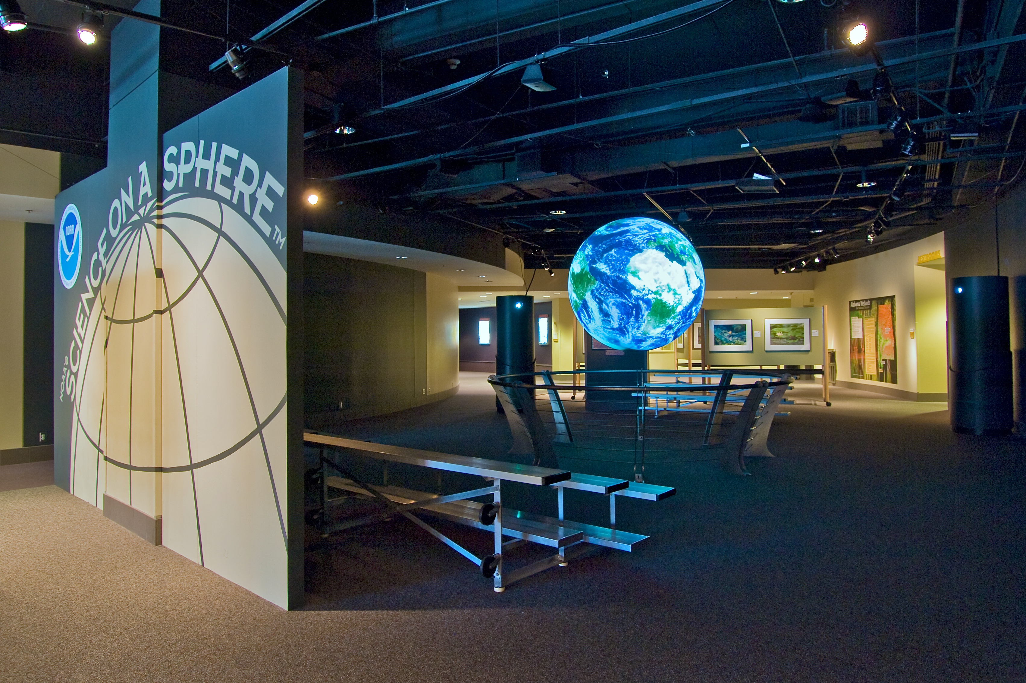 Science On a Sphere hangs in an empty exhibit hall. A partition doubles as a sign reading 'Science On a Sphere' and blocks some of the ambient light coming in from the left