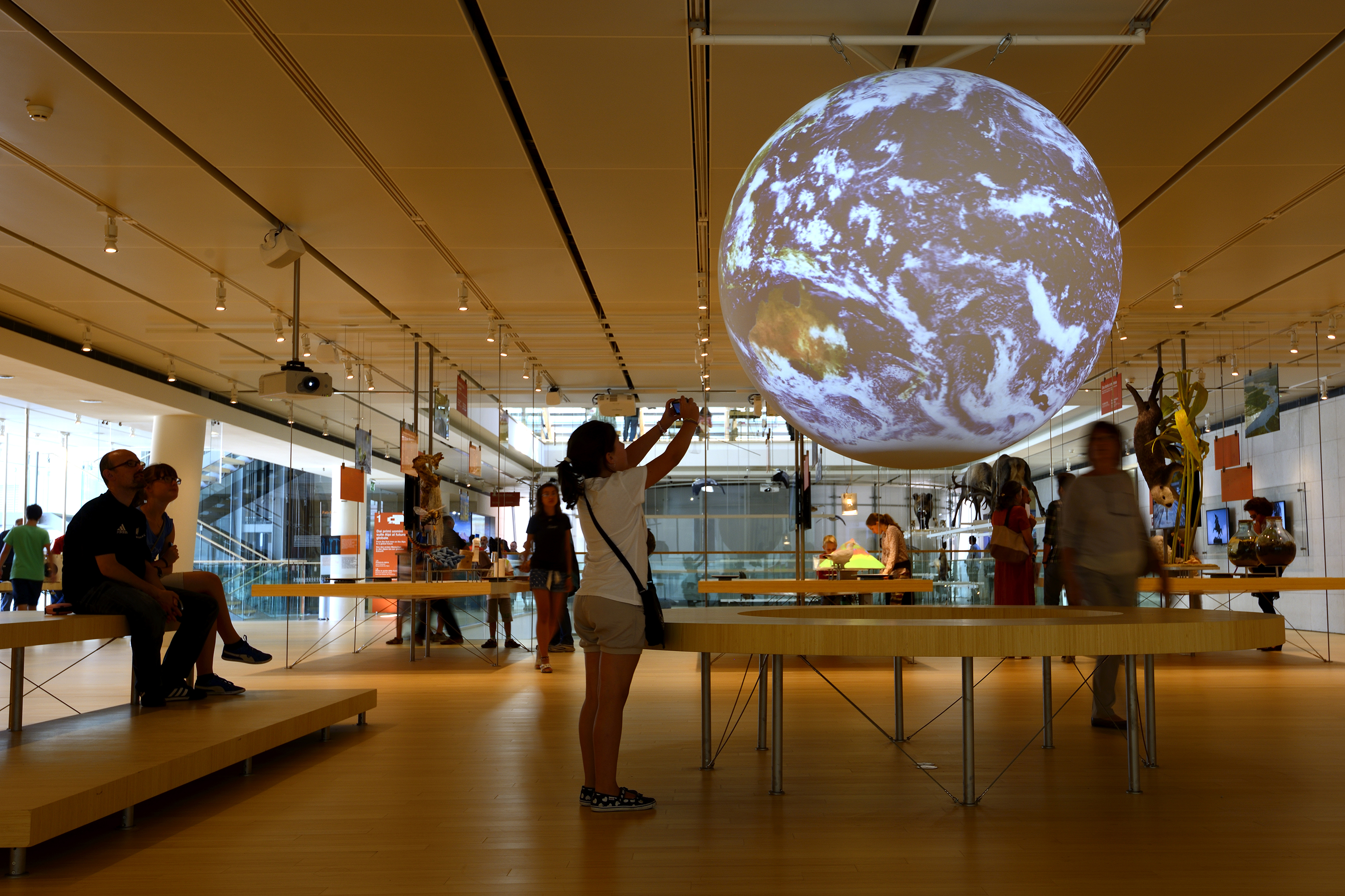 Science On a Sphere displays satellite imagery of Earth in a well-lit room. A visitor leans against the wooden railing surrounding the Sphere and holds out her phone to take a photo. In the background other exhitibts at the museum are visible