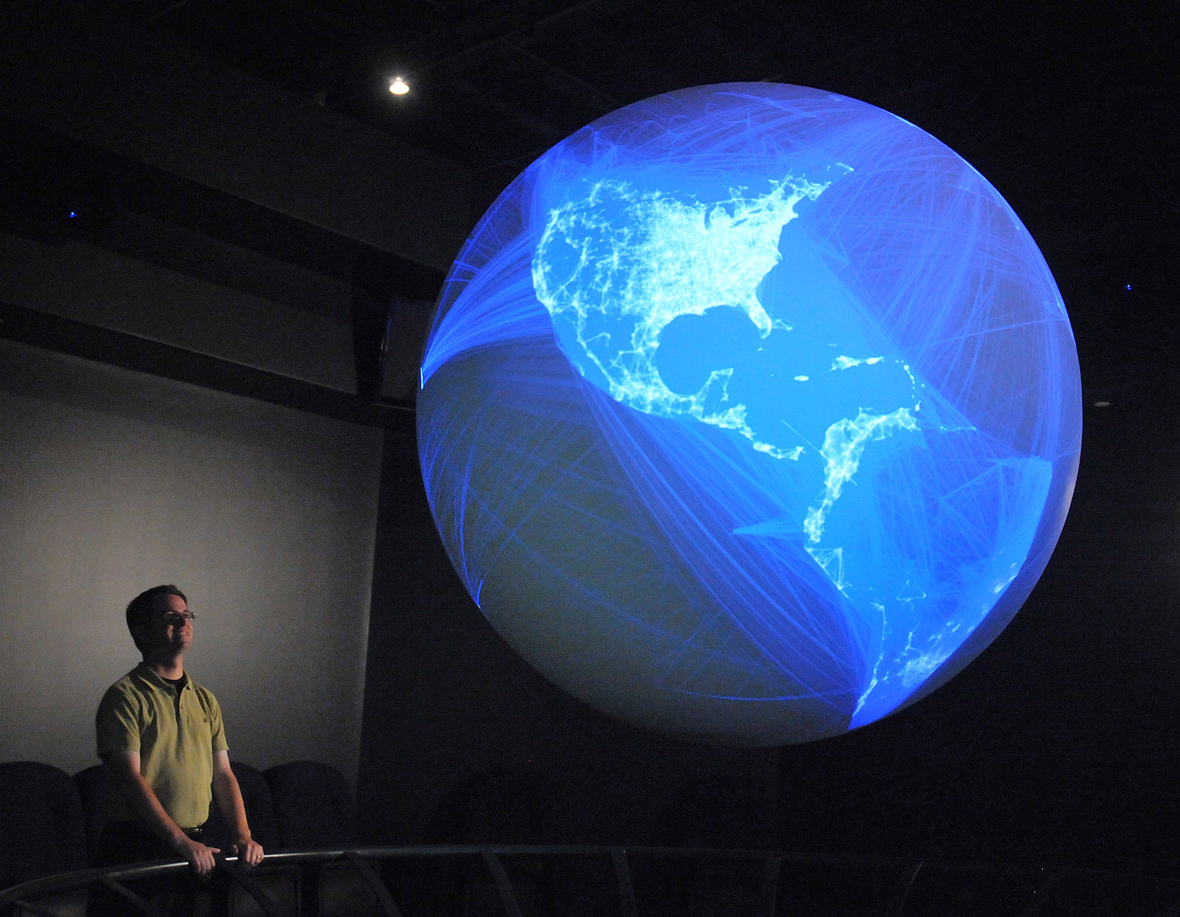A person stands looking at Science On a Sphere displaying thousands of blue lines connecting different cities around the world