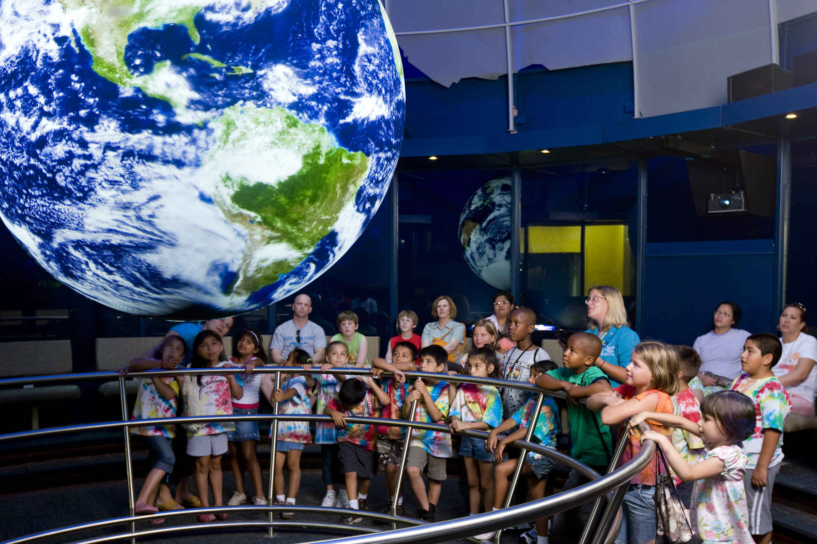 A group of school children crowd around a railing watching Science On a Sphere as a docent stands behind them giving a presentation