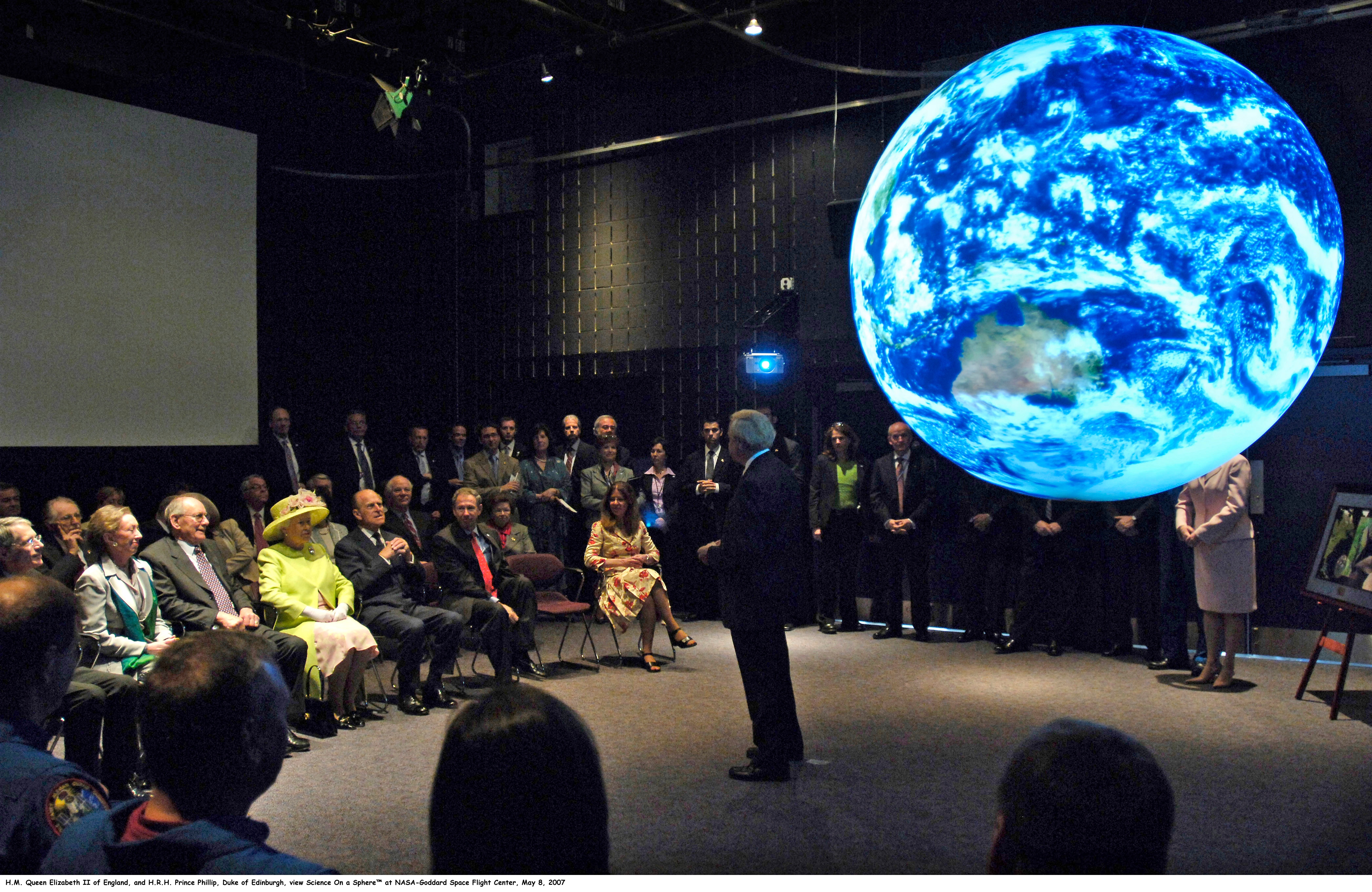 A group of people sit and stand around Science On a Sphere as it displays satellite imagery of Earth in a theater. One man stands in front of the Sphere facing the audience. Queen Elizabeth II is seated in the front row, wearing a lime green jacket and matching hat; seated to her left is Prince Phillip in a dark suit