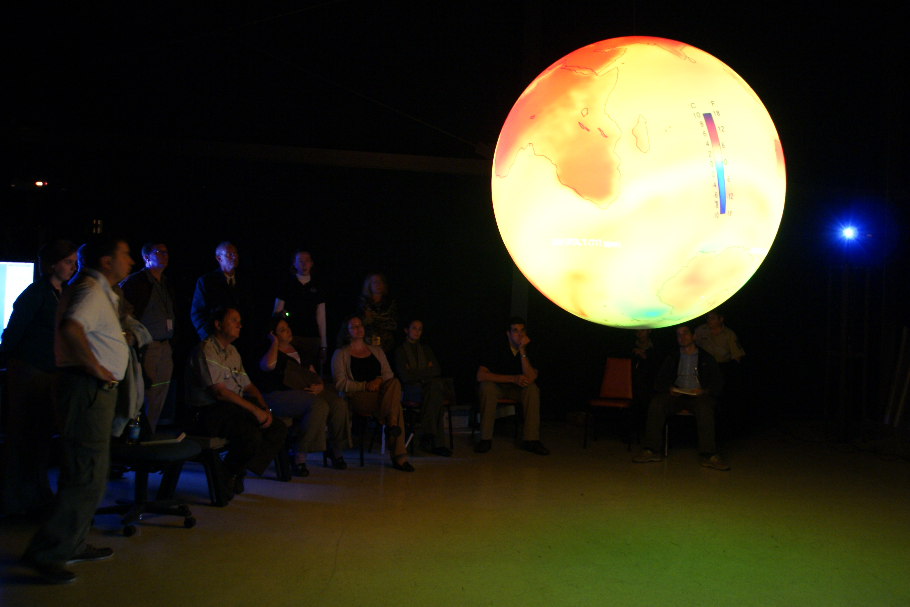 A group of adults stand or sit in a semicircle watching Science On a Sphere displaying global temperature data
