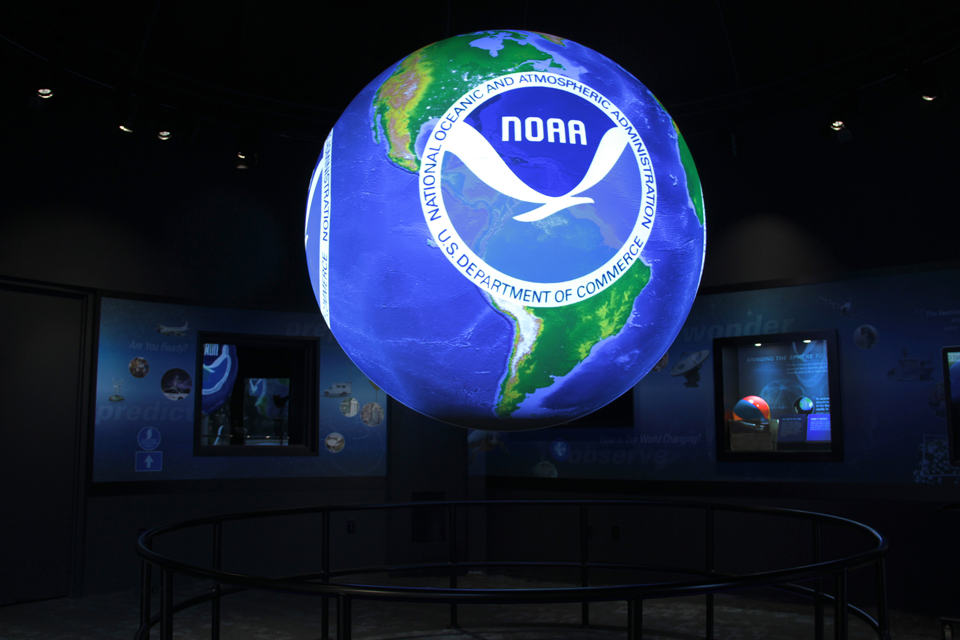 Science On a Sphere displays the NOAA logo atop an image of Earth. In the background, display cases containing other NOAA-related objects are visible